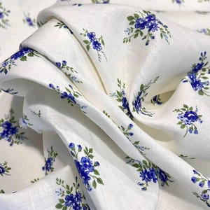Pu&ograve; includere: Tessuto di lino bianco con un piccolo motivo floreale blu. Il tessuto &egrave; piegato e drappeggiato, mostrando la trama del lino.