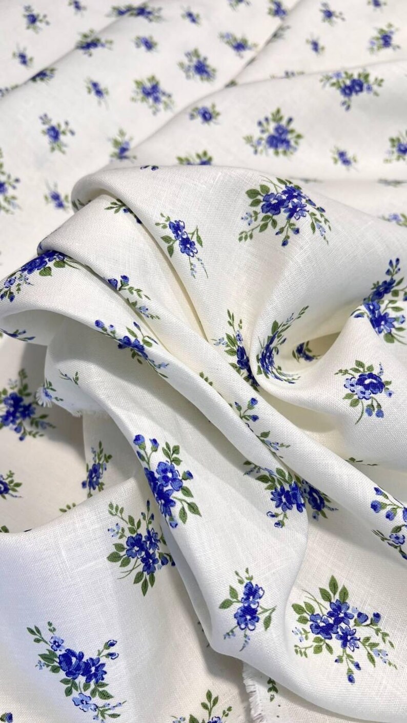 Pu&ograve; includere: Tessuto di lino bianco con un piccolo motivo floreale blu. Il tessuto &egrave; piegato e drappeggiato, mostrando la trama del lino.
