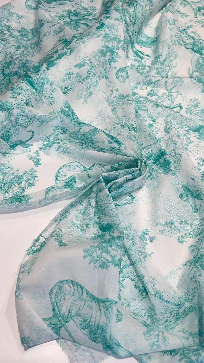 May include: White fabric with a teal toile pattern featuring tigers, trees, and other wildlife. The fabric is folded, showing the detailed design of the forest scene. Ideal for home decor or apparel.