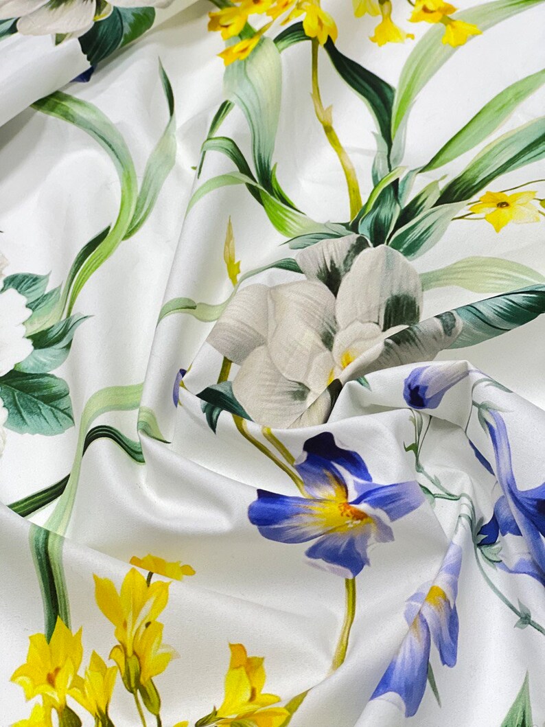 May include: White fabric with a floral print featuring yellow, blue, and white flowers. The fabric has a soft, flowing texture.
