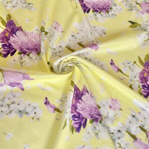 May include: A close-up of a yellow fabric with a floral pattern. The design features large purple and white flowers with green leaves. The fabric is folded, creating shadows and depth, highlighting the texture and the intricate floral details.