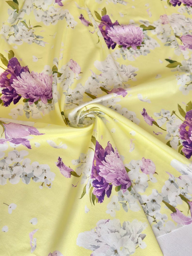 May include: A close-up of a yellow fabric with a floral pattern. The design features large purple and white flowers with green leaves. The fabric is folded, creating shadows and depth, highlighting the texture and the intricate floral details.