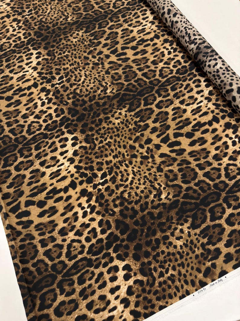 May include: A roll of fabric with a leopard print design. The fabric features a pattern of dark brown spots on a tan and brown background. The fabric is rolled up, showing the pattern on the edge.