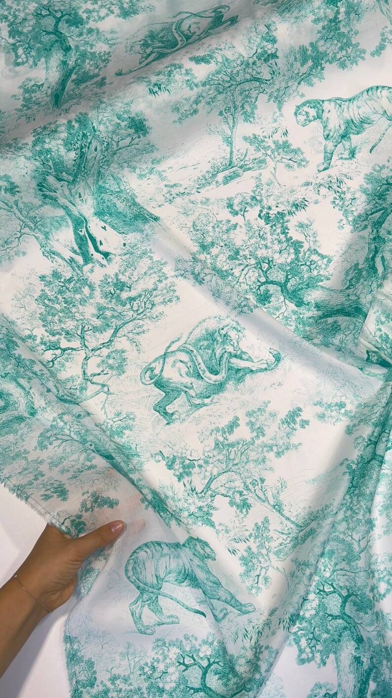 May include: White fabric with a teal toile de Jouy design featuring tigers, lions, and trees. The fabric has a detailed, vintage aesthetic, with the animals and foliage rendered in a classic, illustrative style. The fabric is being held by a hand.