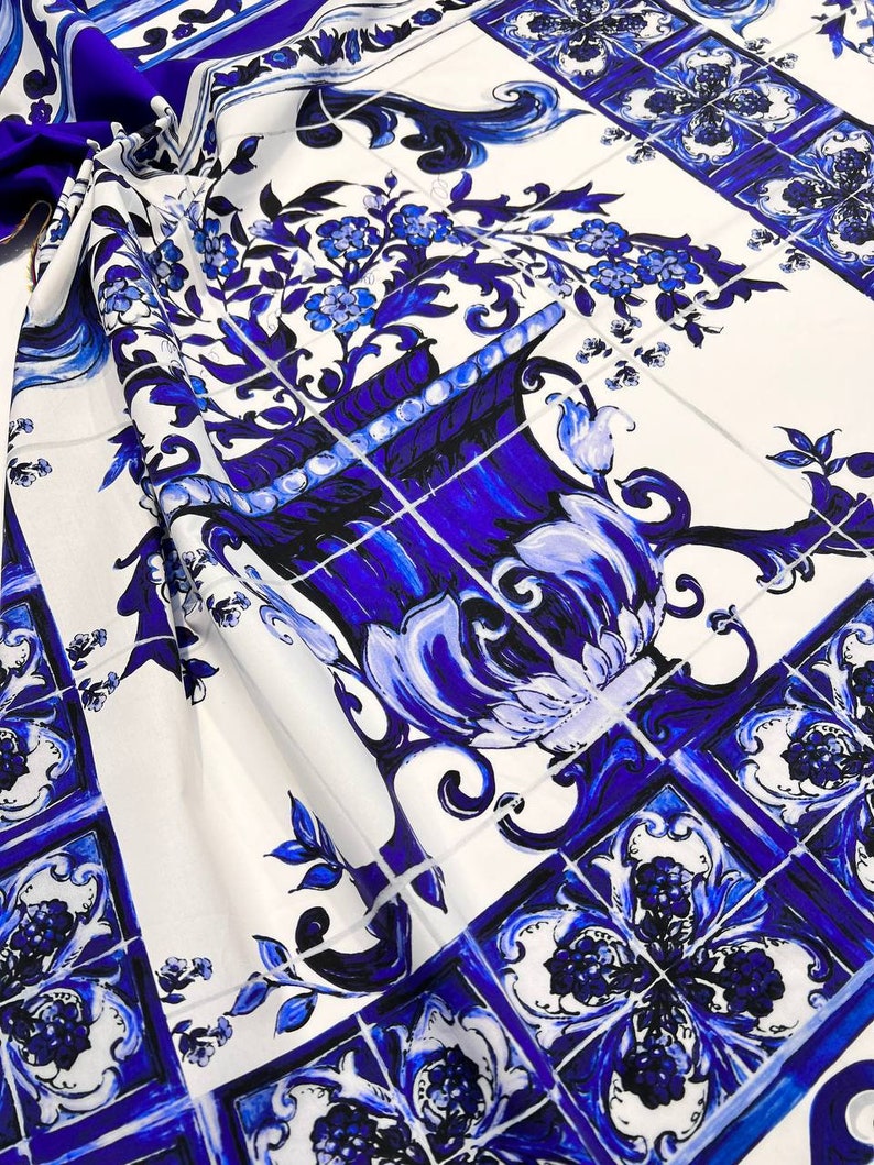 May include: White fabric with a blue and white tile pattern. The pattern features a large blue vase with flowers and intricate designs.