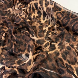 Puede incluir: Una tela fluida con estampado de leopardo. El material presenta manchas marrones y negras sobre un fondo beige. La tela parece ligera y ligeramente brillante. La tela está drapeada.