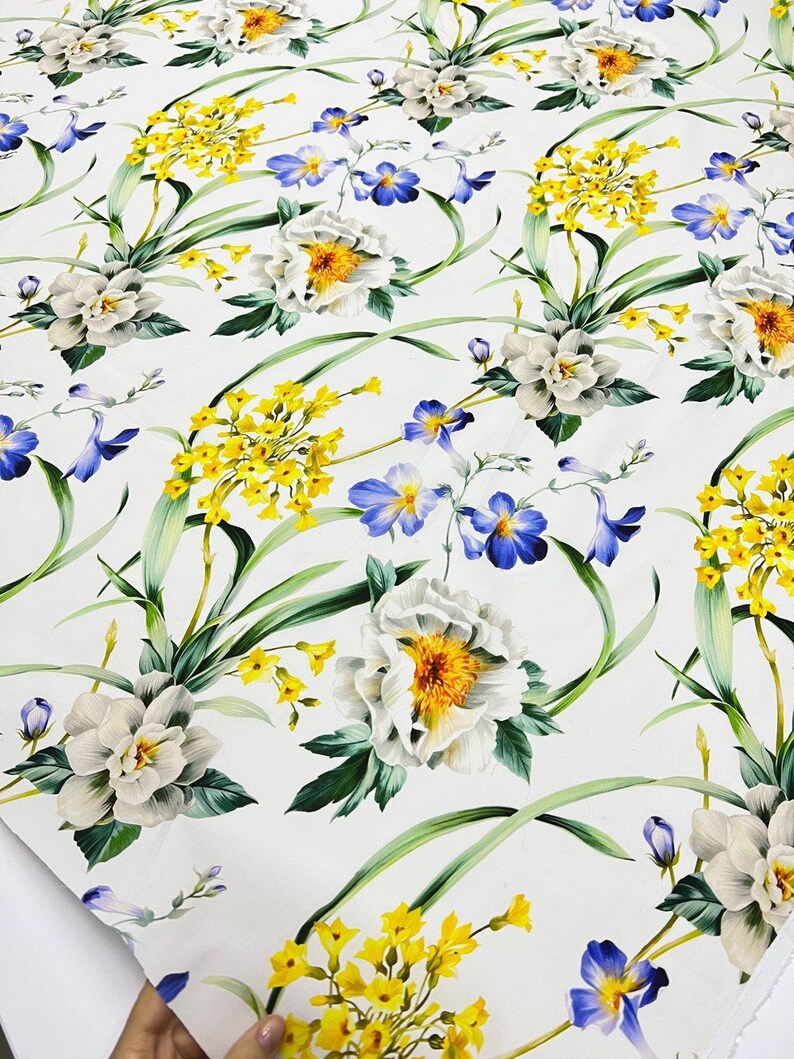 May include: White fabric with a floral print featuring white, yellow, and blue flowers. The flowers are arranged in a repeating pattern.