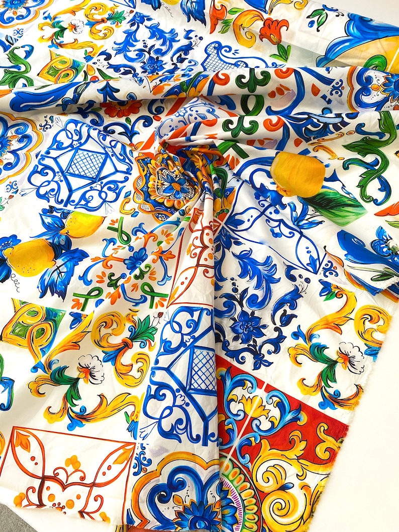 Pu&ograve; includere: Tessuto bianco con un motivo di piastrelle italiane colorate. Il motivo presenta colori blu, gialli, verdi, rossi e bianchi. Il tessuto &egrave; piegato a met&agrave;, mostrando il motivo su entrambi i lati.