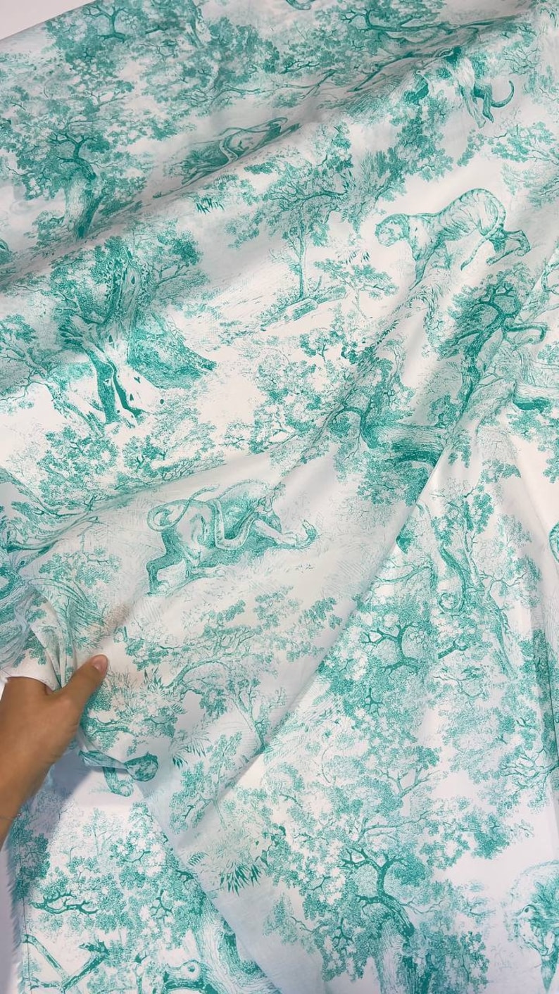 May include: White fabric with a teal toile de Jouy pattern. The design features detailed illustrations of trees, tigers, and other animals in a forest setting. The fabric is folded, showing the pattern's repeat.