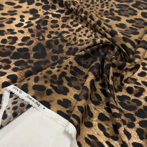 May include: A close-up of a fabric with a leopard print pattern. The fabric is a mix of brown and tan with black spots. The edge of the fabric is white with text that reads "100% Co Made in Italy".
