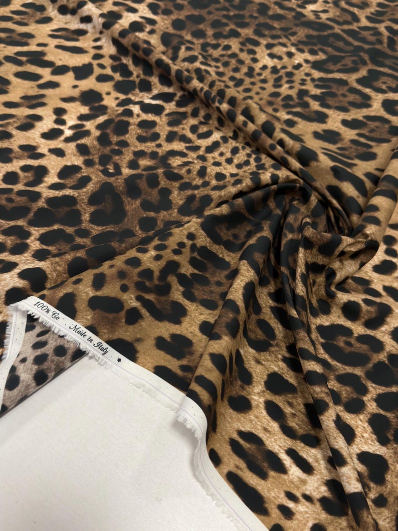 May include: A close-up of a fabric with a leopard print pattern. The fabric is a mix of brown and tan with black spots. The edge of the fabric is white with text that reads "100% Co Made in Italy".