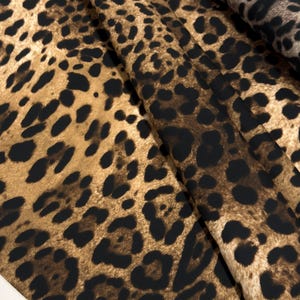 May include: Close-up of a fabric with a leopard print pattern. The fabric features a tan background with dark brown and black spots. The fabric is folded, showing the texture and pattern variations.