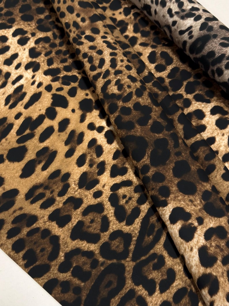 May include: Close-up of a fabric with a leopard print pattern. The fabric features a tan background with dark brown and black spots. The fabric is folded, showing the texture and pattern variations.
