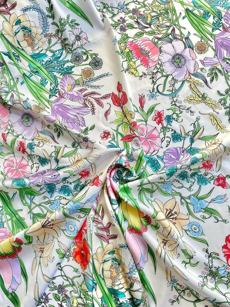 Op de afbeelding: Witte stof met een bloemenprint met verschillende bloemen in tinten roze, paars, geel en rood. De stof is gevouwen en heeft een subtiele glans.