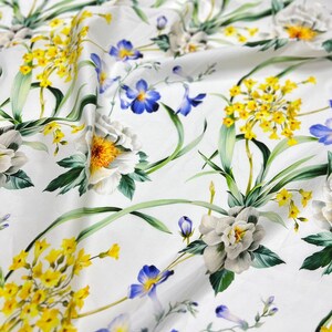 May include: White fabric with a floral print featuring yellow, blue, and white flowers. The fabric has a smooth, silky texture.