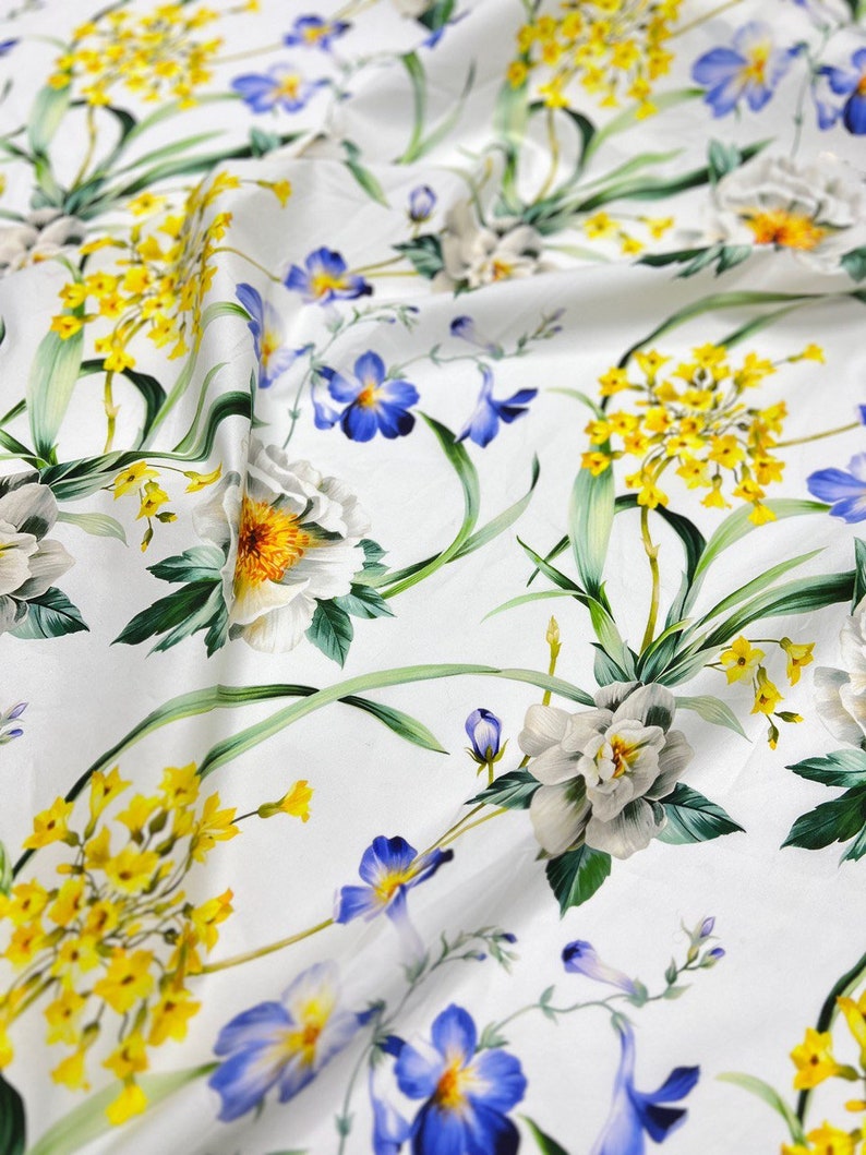 May include: White fabric with a floral print featuring yellow, blue, and white flowers. The fabric has a smooth, silky texture.