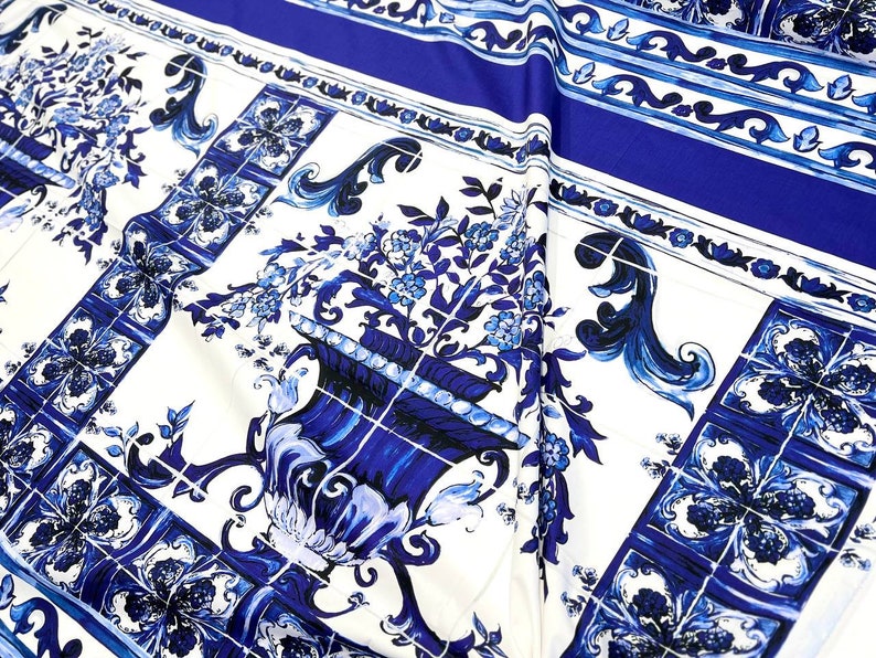 May include: White fabric with a blue and white tile pattern. The pattern features a repeating design of stylized flowers and vases.