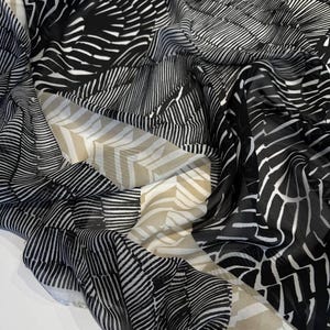 May include: A black and white patterned scarf with a geometric design. The scarf features a variety of patterns, including stripes and curved lines, creating a visually dynamic textile. The scarf is draped, showcasing its flowing fabric.