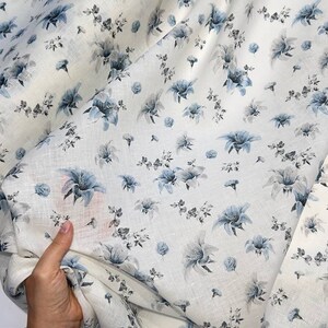 May include: White linen fabric with a blue floral print. The fabric is folded and draped over a hand.