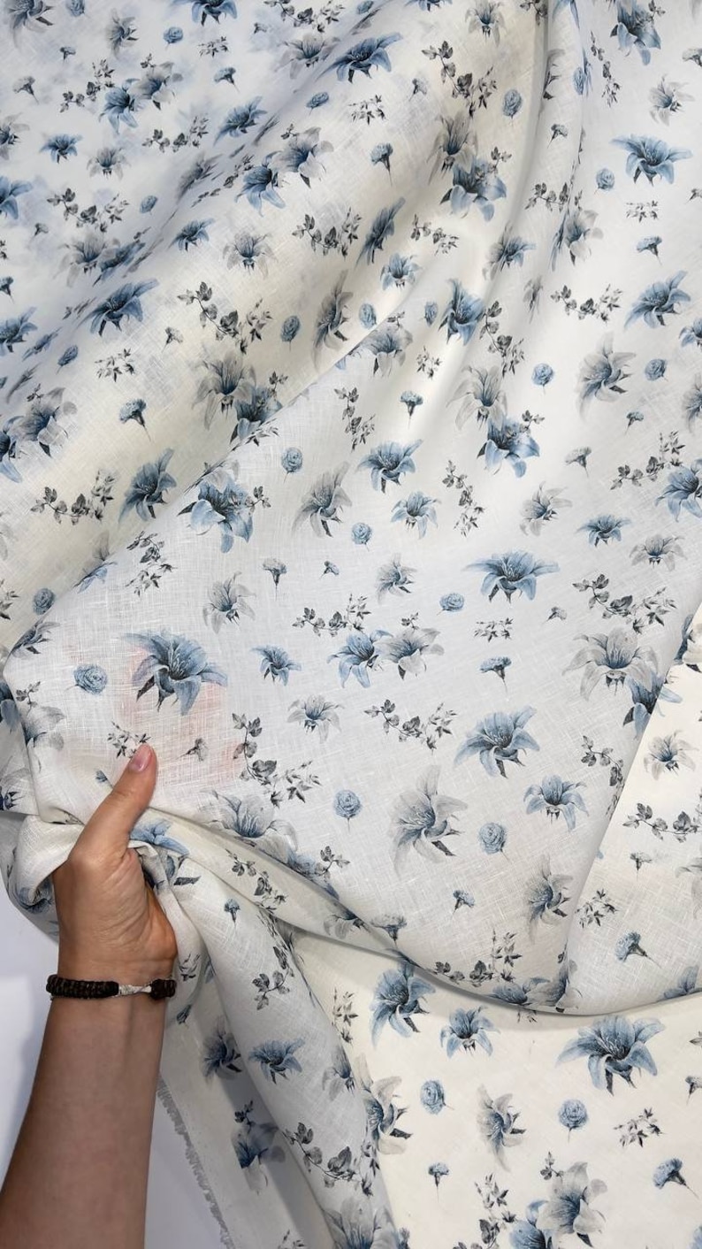 May include: White linen fabric with a blue floral print. The fabric is folded and draped over a hand.