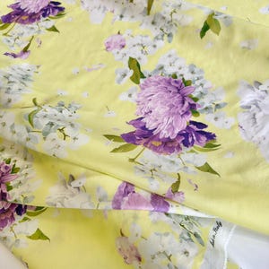 May include: Yellow fabric with a floral pattern featuring large purple and white flowers. The fabric has a soft, flowing appearance, with visible folds and creases. Green leaves accent the floral design, creating a vibrant and colorful textile.