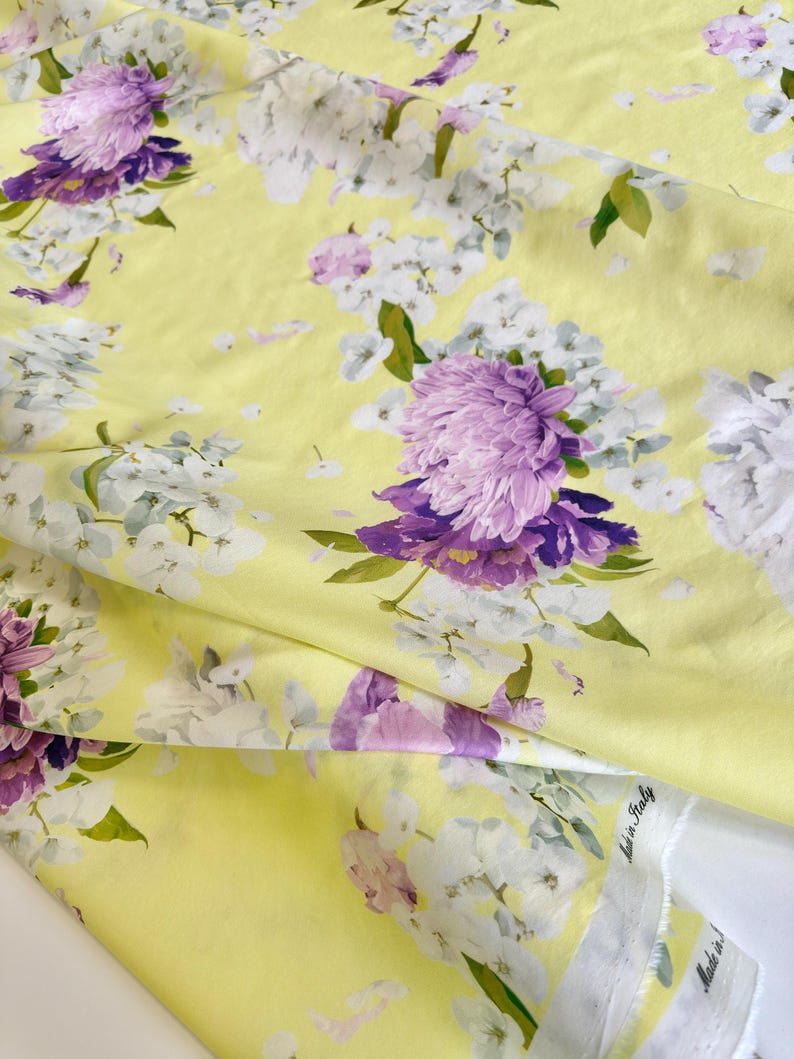 May include: Yellow fabric with a floral pattern featuring large purple and white flowers. The fabric has a soft, flowing appearance, with visible folds and creases. Green leaves accent the floral design, creating a vibrant and colorful textile.