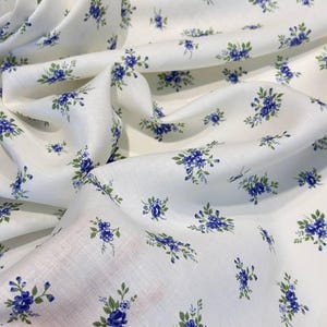 Pu&ograve; includere: Tessuto bianco con una piccola stampa floreale blu. Il tessuto &egrave; piegato e drappeggiato su una mano.