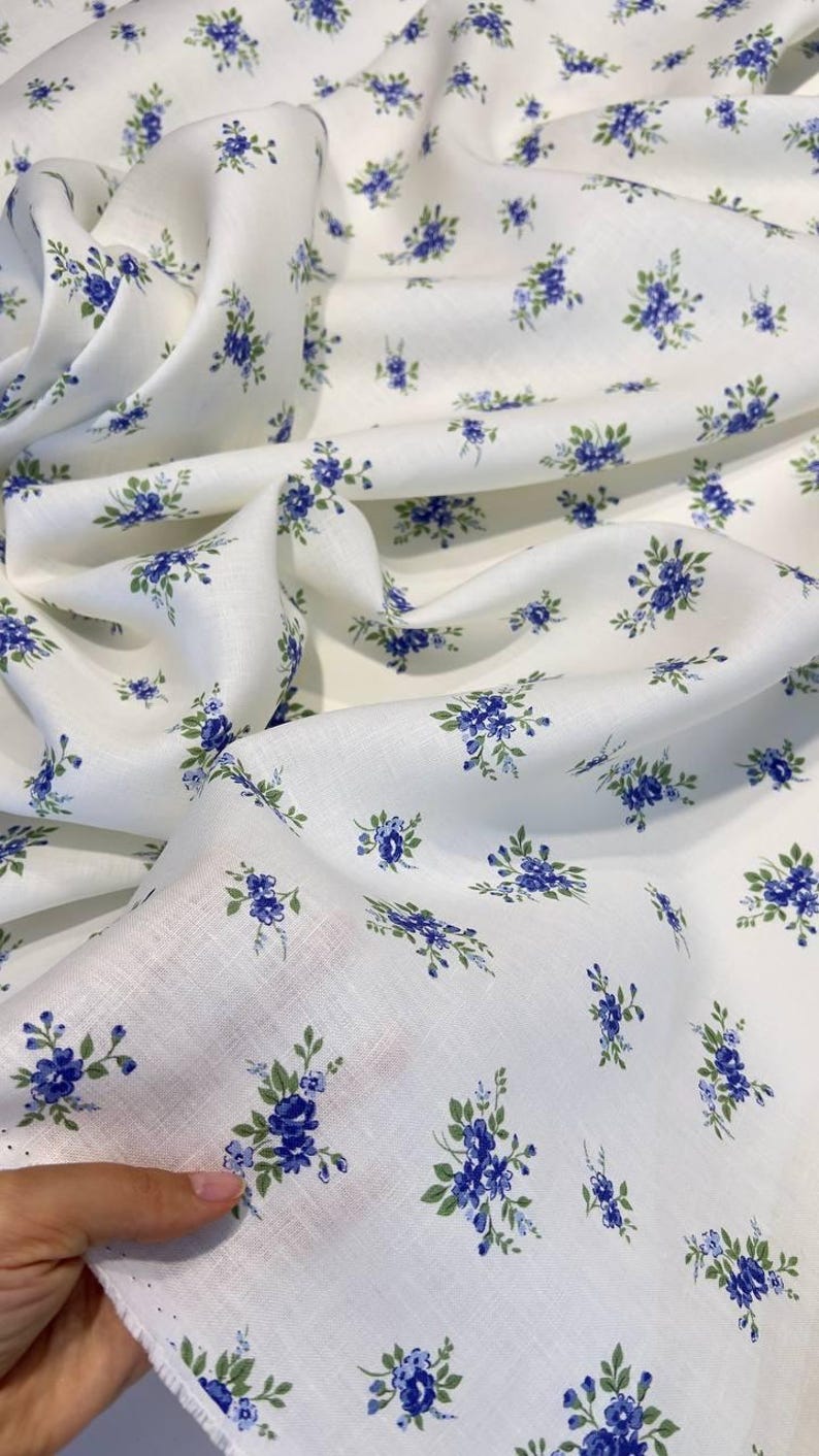 Pu&ograve; includere: Tessuto bianco con una piccola stampa floreale blu. Il tessuto &egrave; piegato e drappeggiato su una mano.