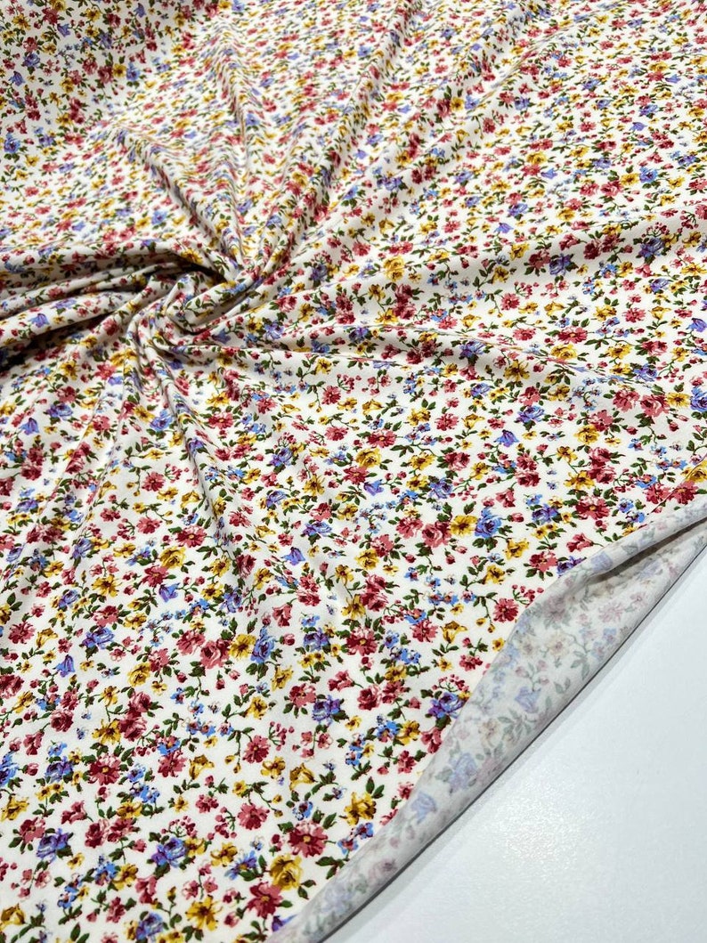 May include: White fabric with a small floral print in shades of red, blue, yellow, and green. The fabric is folded and the underside is visible.