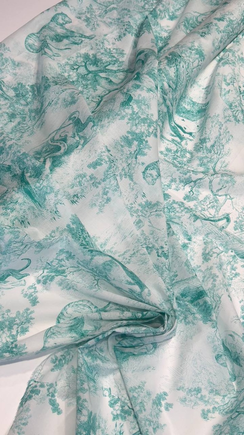 May include: White fabric with a teal green toile de Jouy design. The fabric features a repeating pattern of trees, animals, and figures in a forest setting. The fabric is folded, showing the texture and drape of the material.