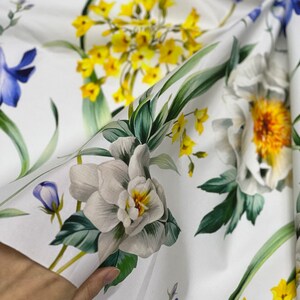 May include: White fabric with a floral print featuring yellow, white, and blue flowers. The fabric is folded in half, and a hand is holding the fabric.
