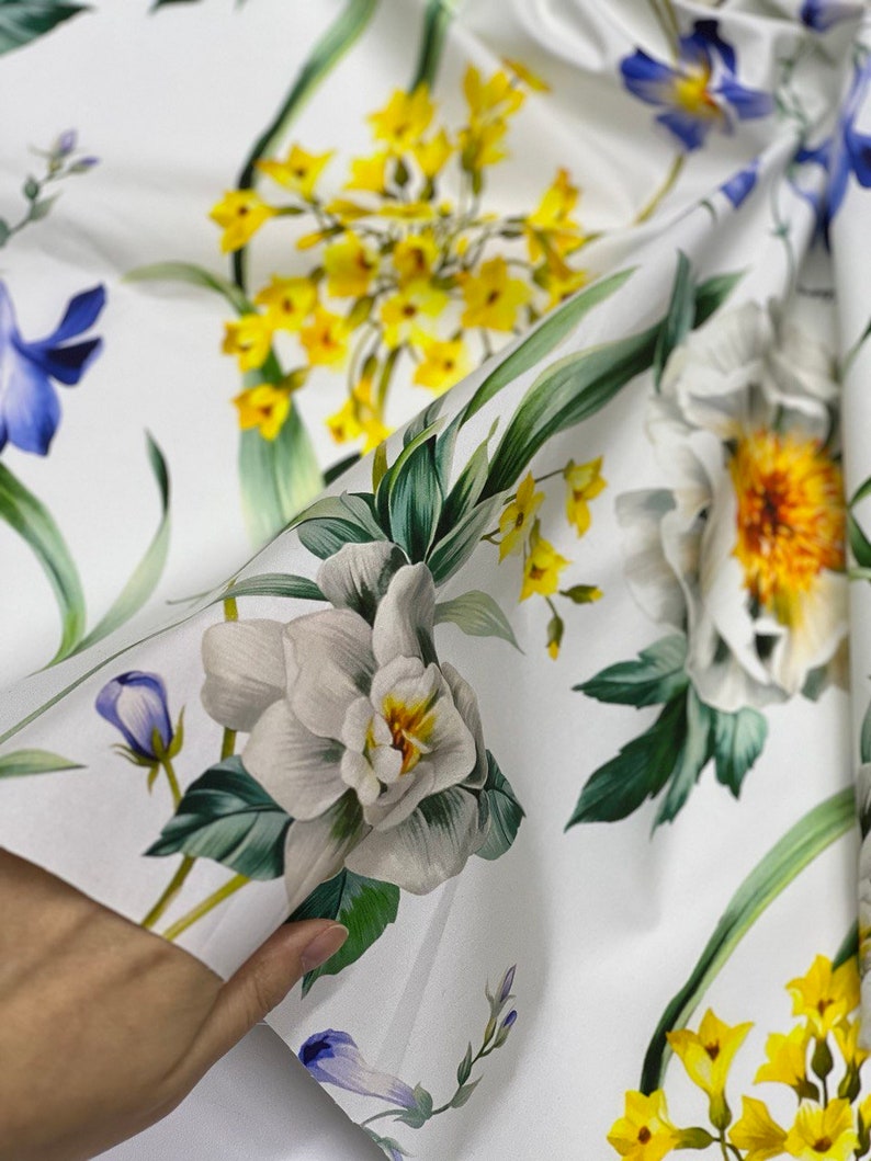 May include: White fabric with a floral print featuring yellow, white, and blue flowers. The fabric is folded in half, and a hand is holding the fabric.