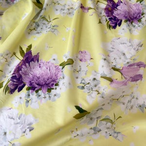 May include: A close-up of a yellow fabric with a floral pattern. The design features large purple and white flowers with green leaves. The fabric is draped, creating soft folds and shadows.