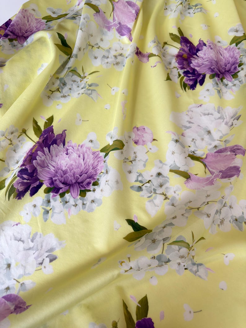 May include: A close-up of a yellow fabric with a floral pattern. The design features large purple and white flowers with green leaves. The fabric is draped, creating soft folds and shadows.