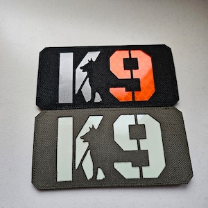 Könnte beinhalten: Zwei schwarze und grüne Patches mit dem Text "K9" in Weiß und einer Silhouette eines Hundes in Weiß. Der obere Patch hat eine orange reflektierende Nummer "9".
