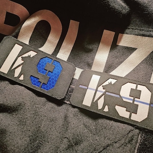 Könnte beinhalten: Zwei schwarze Patches mit weißem Text und blauem Rand. Die Patches zeigen "K9" und eine Silhouette eines Hundes. Ein Patch hat eine blaue Nummer "9" mit einem Wabenmuster.
