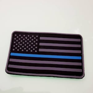Könnte beinhalten: Ein schwarzer und blauer amerikanischer Flaggen-Patch mit einem einzigen blauen Streifen in der Mitte. Die Sterne sind weiß und die Streifen sind grau.