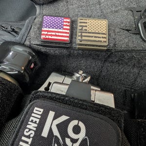 以下が含まれることがあります： アメリカ国旗のデザインが施された2つの正方形のパッチ。1つは紫色の星と赤いストライプ、もう1つはベージュと黒の配色です。画像には、犬のグラフィックとともに「DIENSTHUND」と「K9」というテキストが入った黒いパッチも表示されています。