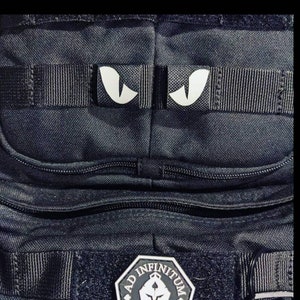 May include: Black tactical backpack with a zippered pocket and a patch that says "AD INFINITUM" with a stylized design. The backpack has two white eye-shaped patches on the top strap.