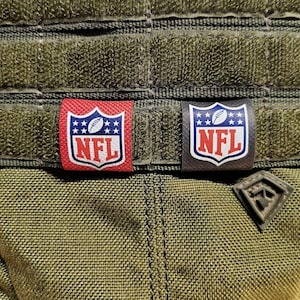 Peut inclure: Deux patchs NFL, l'un rouge et l'autre noir, sont fixés à un gilet tactique vert. Les patchs présentent le logo NFL et un ballon de football.