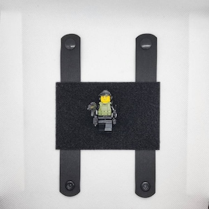 Könnte beinhalten: Schwarze taktische Ausrüstung mit einer Lego-Figur eines Soldaten. Die Ausrüstung hat einen rechteckigen schwarzen Aufnäher mit zwei schwarzen Riemen und Druckknöpfen. Der Lego-Soldat trägt einen Helm und militärische Ausrüstung.