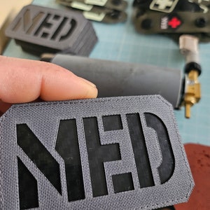 Könnte beinhalten: Ein grauer Stoff-Patch mit dem Wort "MED" ausgeschnitten in Schwarz. Der Patch hat eine strukturierte Oberfläche und ist zum Annähen an Kleidung oder Ausrüstung gedacht.
