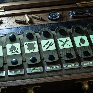 May include: A wooden storage box containing seven olive green fabric straps with glow-in-the-dark icons representing power, fire, food, cutlery, tools, water, and first aid, labeled with German text including Strom, Feuer, Verpflegung, and Erste Hilfe.
