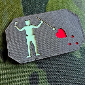 Könnte beinhalten: Ein grauer Stoff-Patch mit einer weißen Silhouette einer Person, die einen Speer hält, der ein rotes Herz durchbohrt. Unter dem Herzen befinden sich drei rote Punkte.