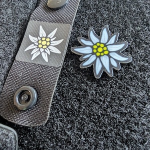 Könnte beinhalten: Ein schwarzer Riemen mit Druckknopfverschluss und einem kleinen Aufnäher mit einem weißen und gelben Blumendesign. Ein separater, ähnlicher blütenförmiger Aufnäher mit blauen Blütenblättern und einem gelben Zentrum ist ebenfalls sichtbar.
