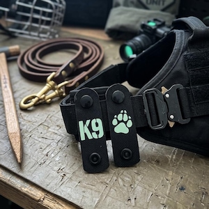 Peut inclure: Deux écussons tactiques en nylon noir avec boutons-pression fixés sur un harnais. Un écusson porte le texte K9 en vert clair, et l'autre affiche une icône de patte en vert clair. Le harnais comprend une boucle en métal noir et des sangles.