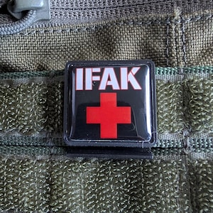 Könnte beinhalten: Ein schwarz-weißes Patch mit dem Text "IFAK" in weißen Buchstaben und einem roten Kreuz in der Mitte. Das Patch ist an einem grünen Molle-Panel befestigt.