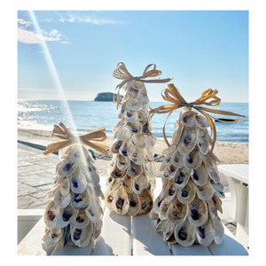 May include: Three decorative seashell Christmas trees with natural raffia bows. The trees are arranged in a row on a white surface, with a beach and ocean in the background. The trees are made of oyster shells.