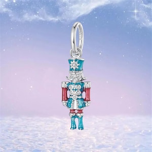 May include: A silver charm pendant in the shape of a toy soldier nutcracker. The nutcracker is painted in blue, red, and silver, with a star on the hat. The charm has a loop at the top for attaching to a bracelet or necklace. The background is a soft blue with white sparkles.