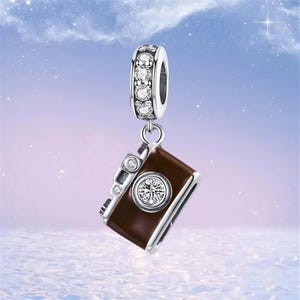 May include: A silver charm with a brown camera pendant. The camera has a round, clear gem in the center and smaller gems on the side. The top of the charm has a ring with clear gems. The background is a soft blue and purple.